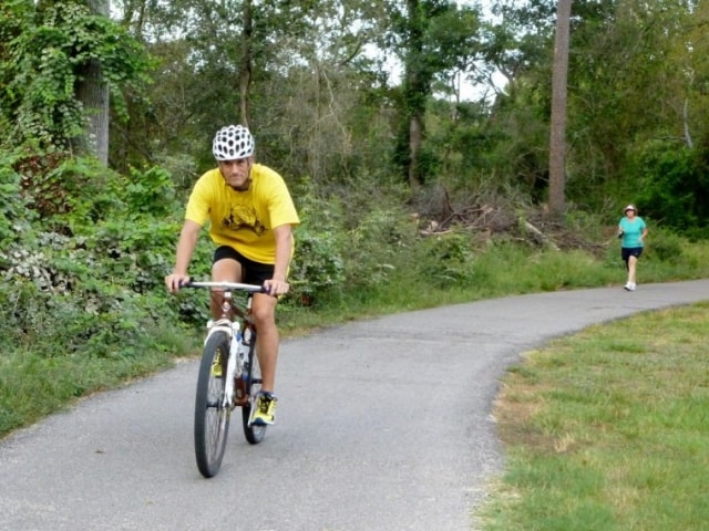 Cyclist in a yellow shirt and helmet riding the Cypress Creek greenway trail with a runner behind him at CreekFest