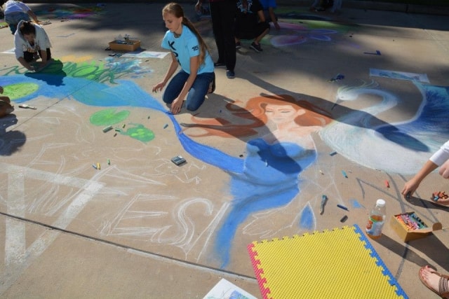 Student chalk artist creating a large mermaid mural on pavement at CreekFest