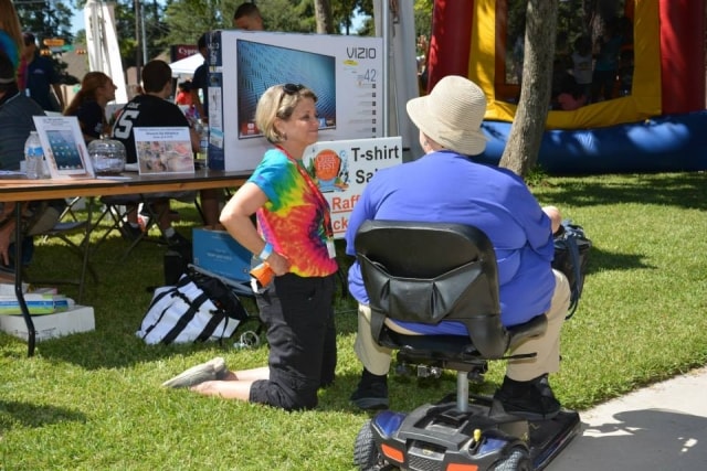 Volunteer in tie-dye shirt kneeling to speak with an attendee on a mobility scooter at the CreekFest t-shirt sale booth