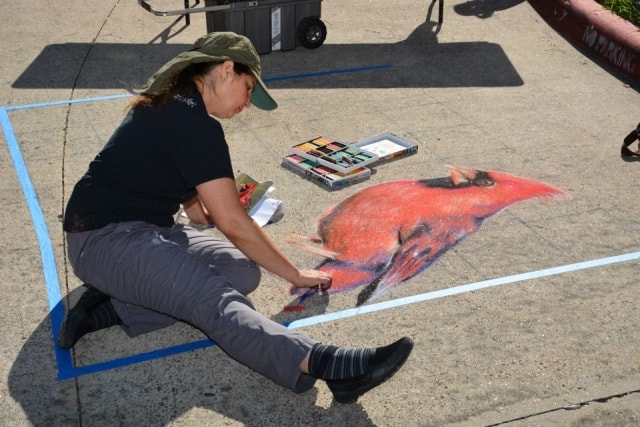 Chalk artist kneeling to paint a detailed red cardinal on the pavement at CreekFest Houston
